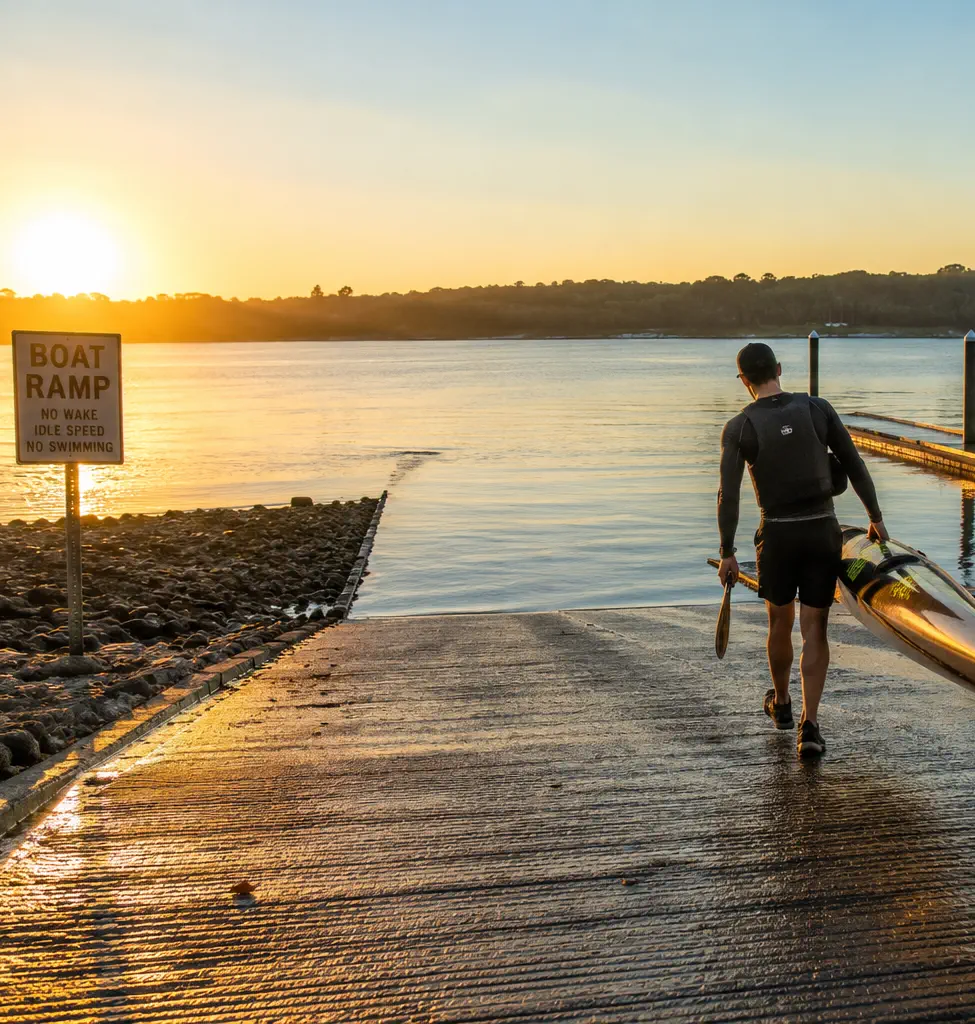 Person carrying a kayak down a public boat ramp before launching on a calm lake